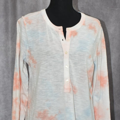 A long-sleeve henley shirt in a light heather gray fabric with soft orange and light blue tie-dye washes, displayed on a black mannequin torso against a gray textured backdrop.