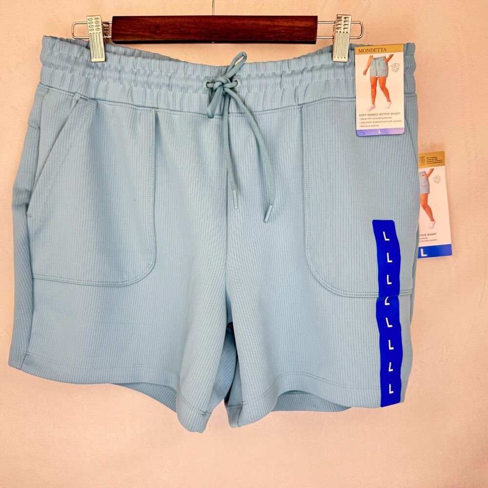 Light blue ribbed athletic shorts with a drawstring waist, side pockets, and a blue vertical stripe with size letters on the left leg, hanging on a wooden hanger against a beige wall.