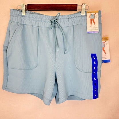 Light blue ribbed athletic shorts with a drawstring waist, side pockets, and a blue vertical stripe with size letters on the left leg, hanging on a wooden hanger against a beige wall.