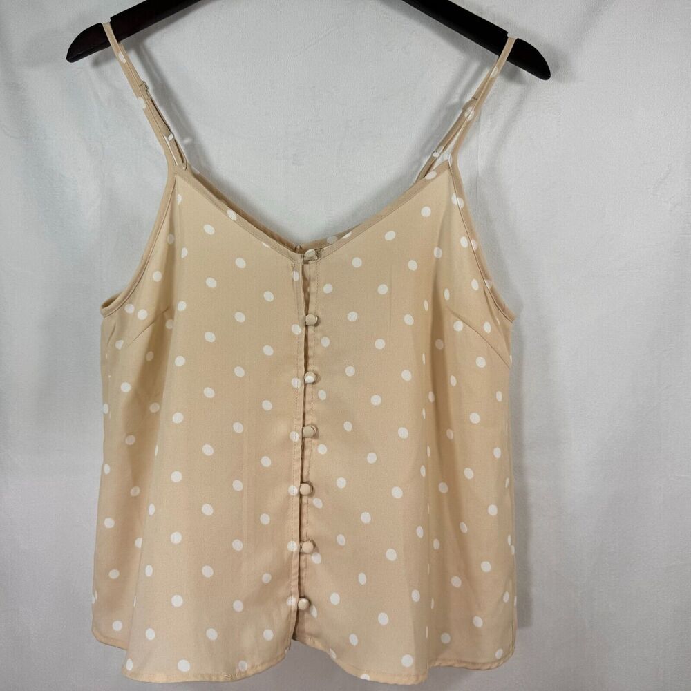 A beige camisole top with white polka dots, featuring a V-neckline, thin shoulder straps, and a decorative button-down front, hanging on a black hanger against a light gray fabric backdrop.