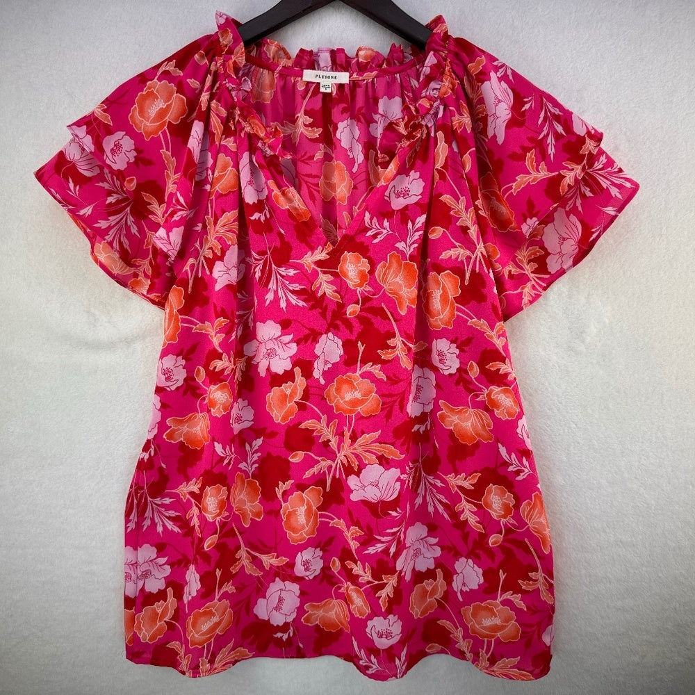A women's short-sleeve blouse with a vibrant pink, orange, and red floral print, hanging on a black hanger against a textured off-white wall.