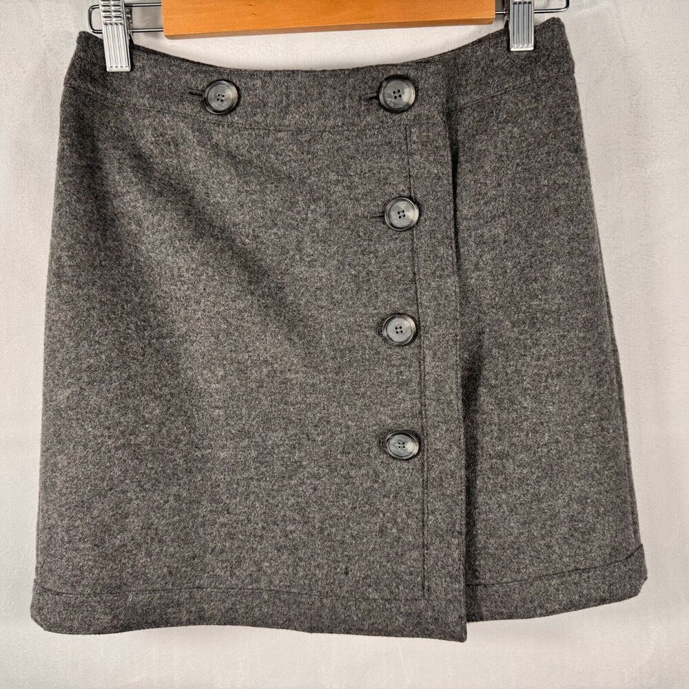 A gray textured wool skirt with a button-front wrap design, hanging on a wooden hanger against a light fabric backdrop.