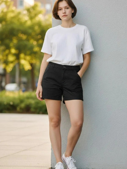 A woman wearing a white crewneck t-shirt and black shorts, leaning against a light gray concrete wall outdoors.