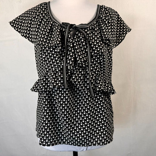 Black and white polka dot blouse with short flutter sleeves, a ruffled neckline, and a front tie detail, displayed on a dress form against a light fabric backdrop.