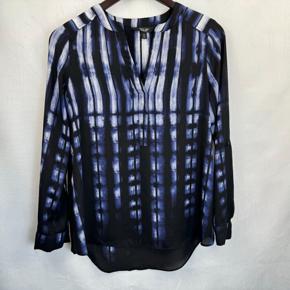 A long-sleeve blouse with a blue and black vertical tie-dye pattern, hanging on a wooden hanger against a white fabric background.