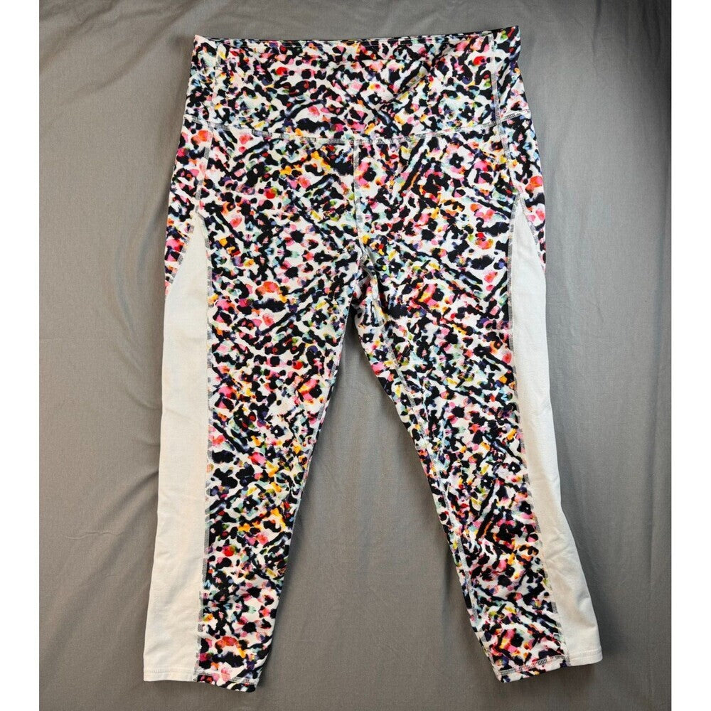 A pair of women's athletic leggings laid flat on a gray fabric background. The leggings feature a dense multicolor abstract splatter pattern in black, white, pink, orange, and yellow, with solid white side panels.