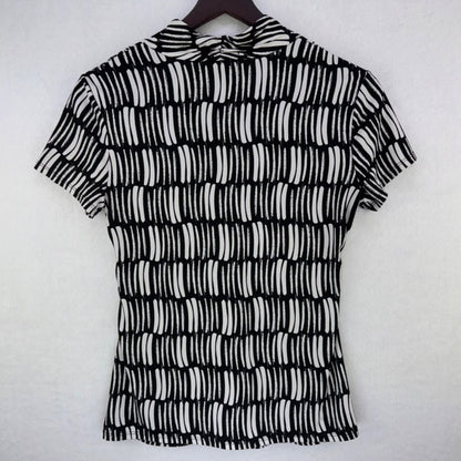 A short-sleeve collared blouse with a black and white abstract wavy stripe pattern, hanging on a dark wooden hanger against a light gray textured wall.