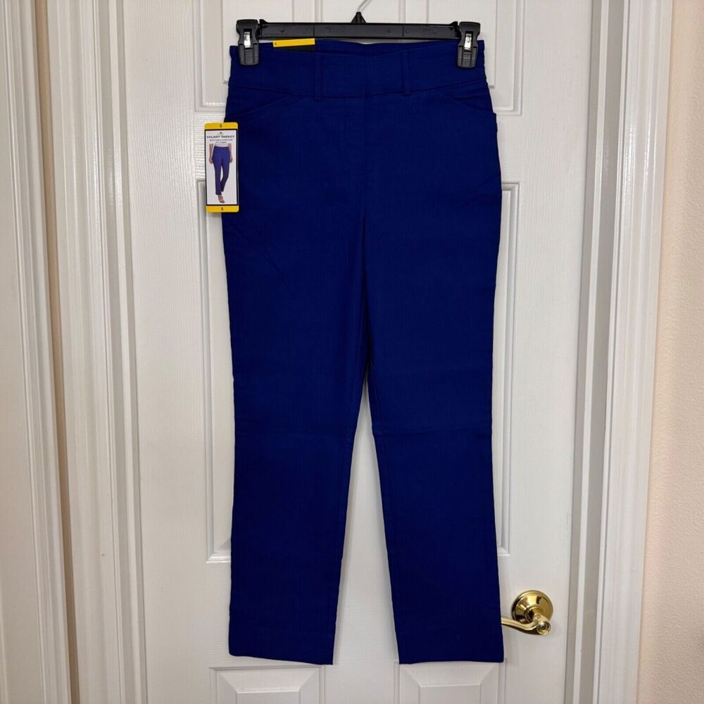 A pair of women's royal blue cropped pants hanging on a black hanger against a white paneled door. The pants are shown with a paper tag attached near the waistband.