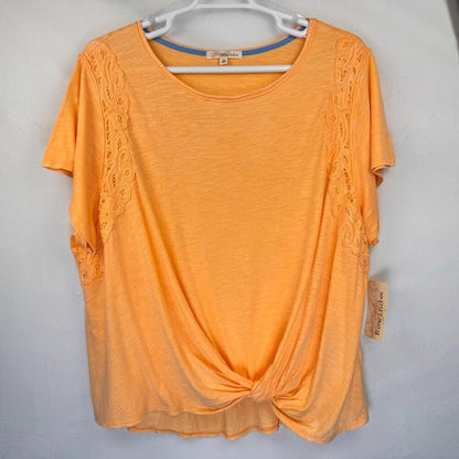 An orange short-sleeve knit top with lace trim on the sides, hanging on a white hanger against a light gray wall. A brand tag is visible on the right side.