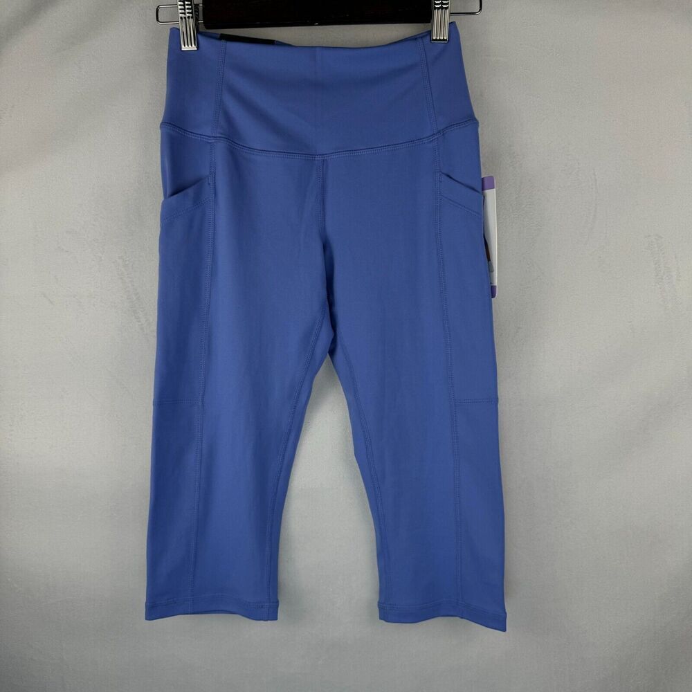 Blue high-waisted cropped leggings with side pockets, hanging on a black hanger against a light gray textured background.