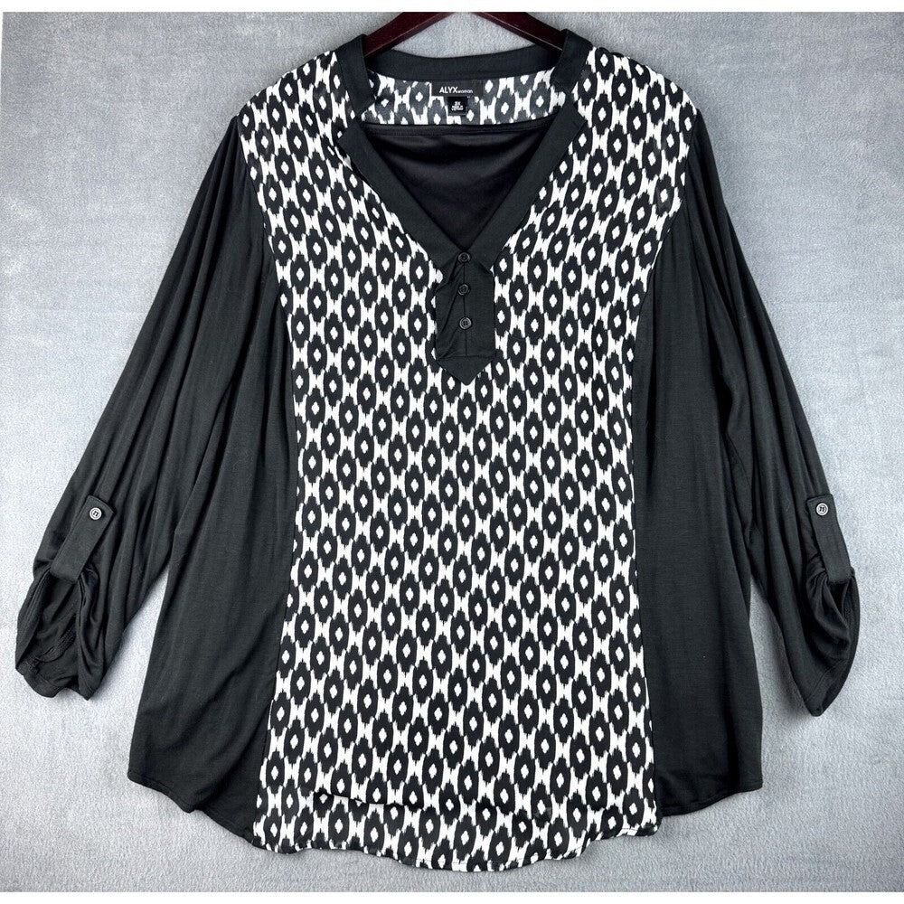 A women's long-sleeve blouse with a black and white ikat pattern on the front panel and solid black sleeves, shown hanging on a dark hanger against a gray textured background.