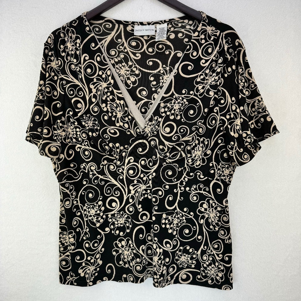 A short-sleeve V-neck blouse with a black background and a beige floral and scroll pattern, hanging on a dark hanger against a light textured wall.