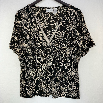 A short-sleeve V-neck blouse with a black background and a beige floral and scroll pattern, hanging on a dark hanger against a light textured wall.