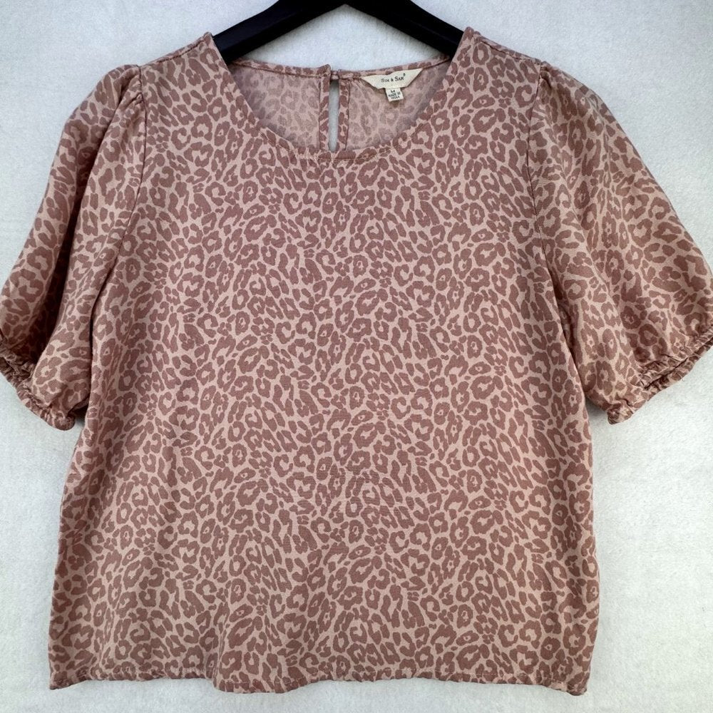 A short-sleeve blouse with an allover leopard print pattern in dusty rose and beige, hanging on a black hanger against a light gray textured background.