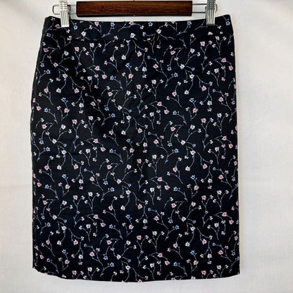 A black skirt with a delicate pink and blue floral vine pattern, hanging on a wooden hanger against a white background.