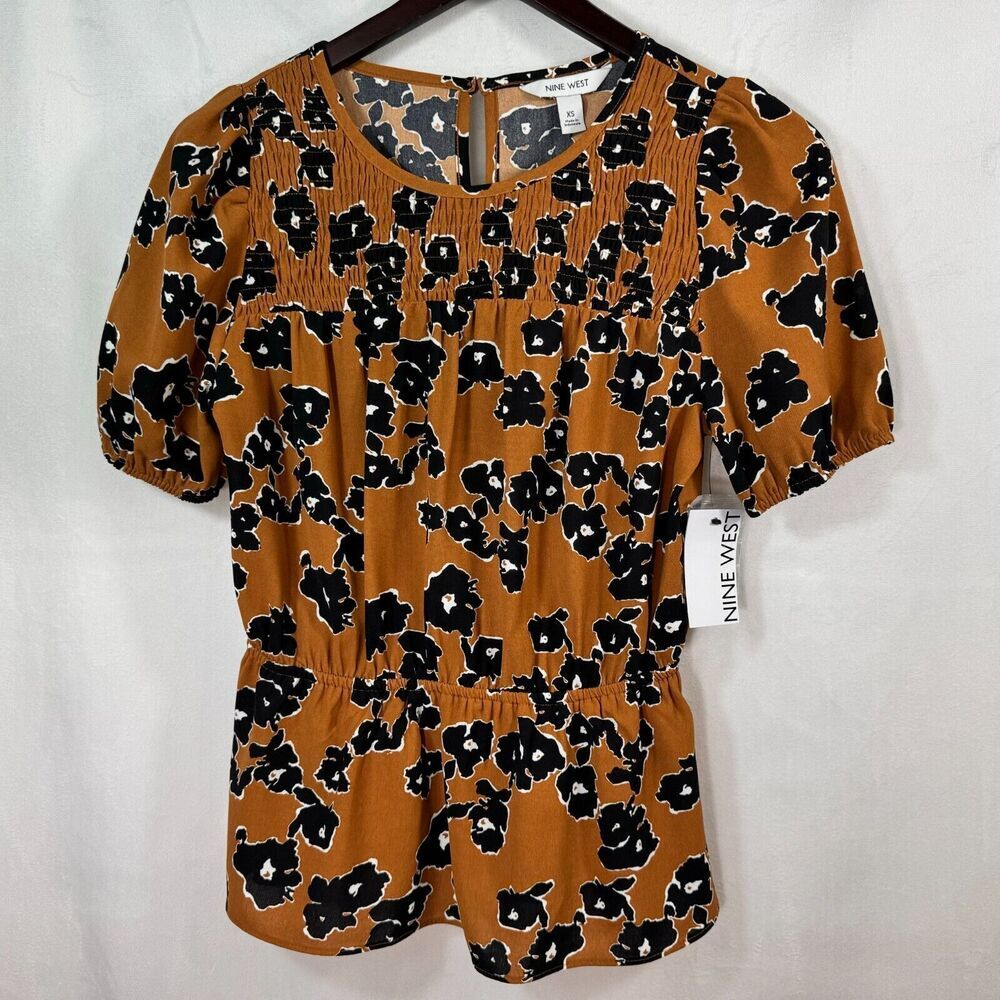 A short-sleeve smocked blouse in a warm brown-orange color with a black and white floral pattern, hanging on a dark wooden hanger against a white fabric backdrop. A 'NINE VENTURES' brand tag is visible on the right side.