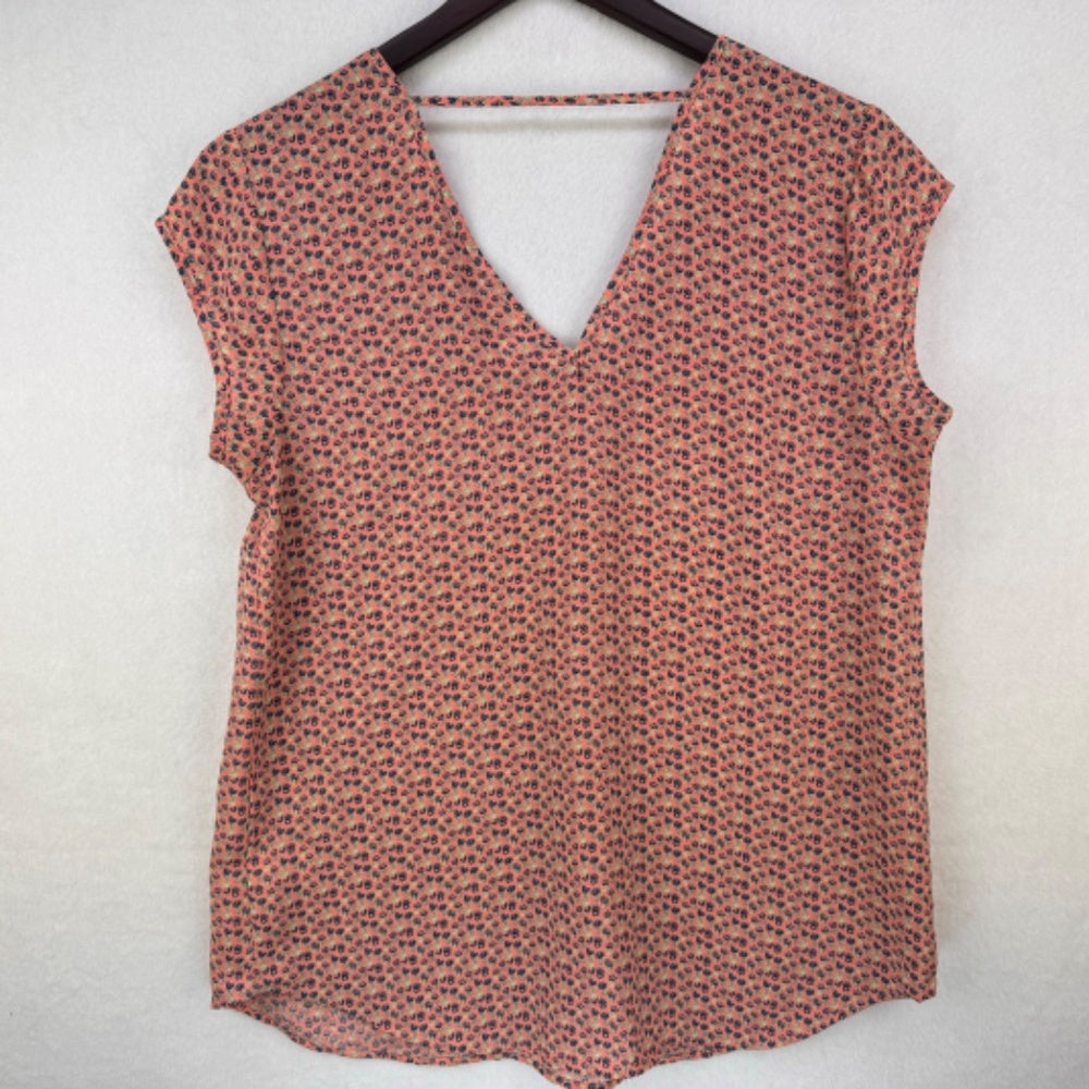 A sleeveless V-neck top with a small-scale red and blue floral pattern on a warm pinkish-beige background, hanging on a black hanger against a textured off-white wall.