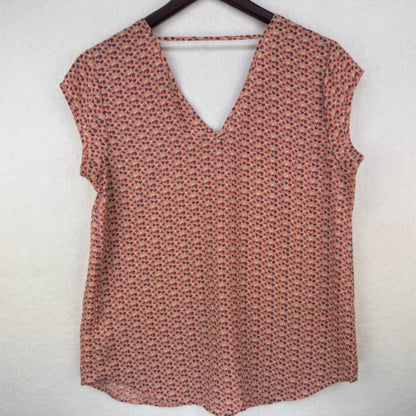 A sleeveless V-neck top with a small-scale red and blue floral pattern on a warm pinkish-beige background, hanging on a black hanger against a textured off-white wall.
