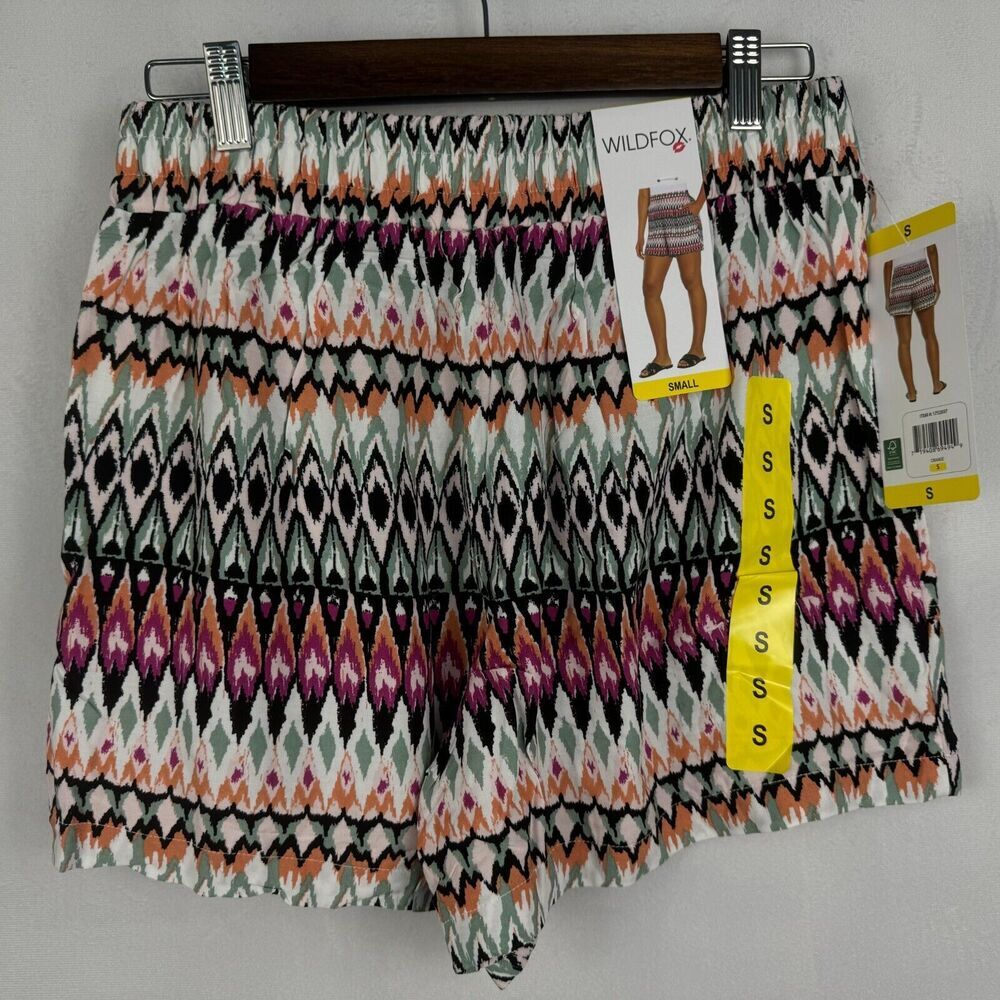 A pair of women's shorts with an intricate geometric ikat pattern in black, white, orange, and purple, hanging on a wooden hanger with retail tags.