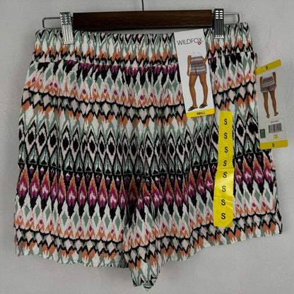 A pair of women's shorts with an intricate geometric ikat pattern in black, white, orange, and purple, hanging on a wooden hanger with retail tags.