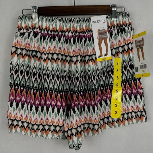 A pair of women's shorts with an intricate geometric ikat pattern in black, white, orange, and purple, hanging on a wooden hanger with retail tags.