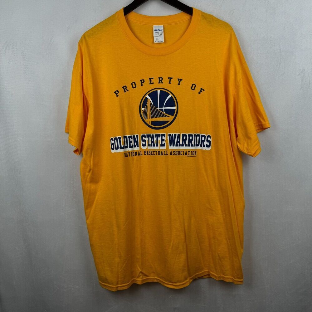 A yellow Golden State Warriors t-shirt hanging on a black hanger against a gray wall. The front of the shirt features a navy and gold graphic with the text 'PROPERTY OF GOLDEN STATE WARRIORS' and the NBA logo.