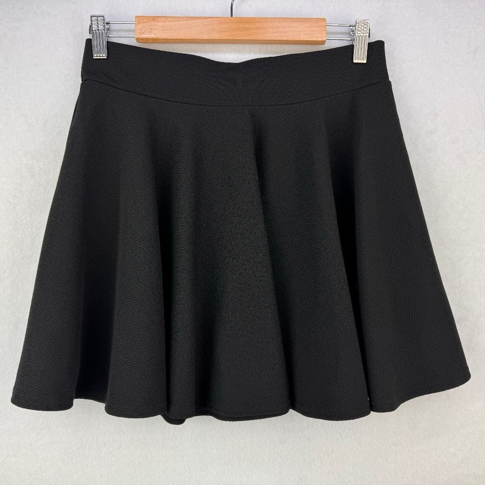 A black flared mini skirt with a textured fabric, hanging from a wooden hanger with silver metal clips against a light gray wall.