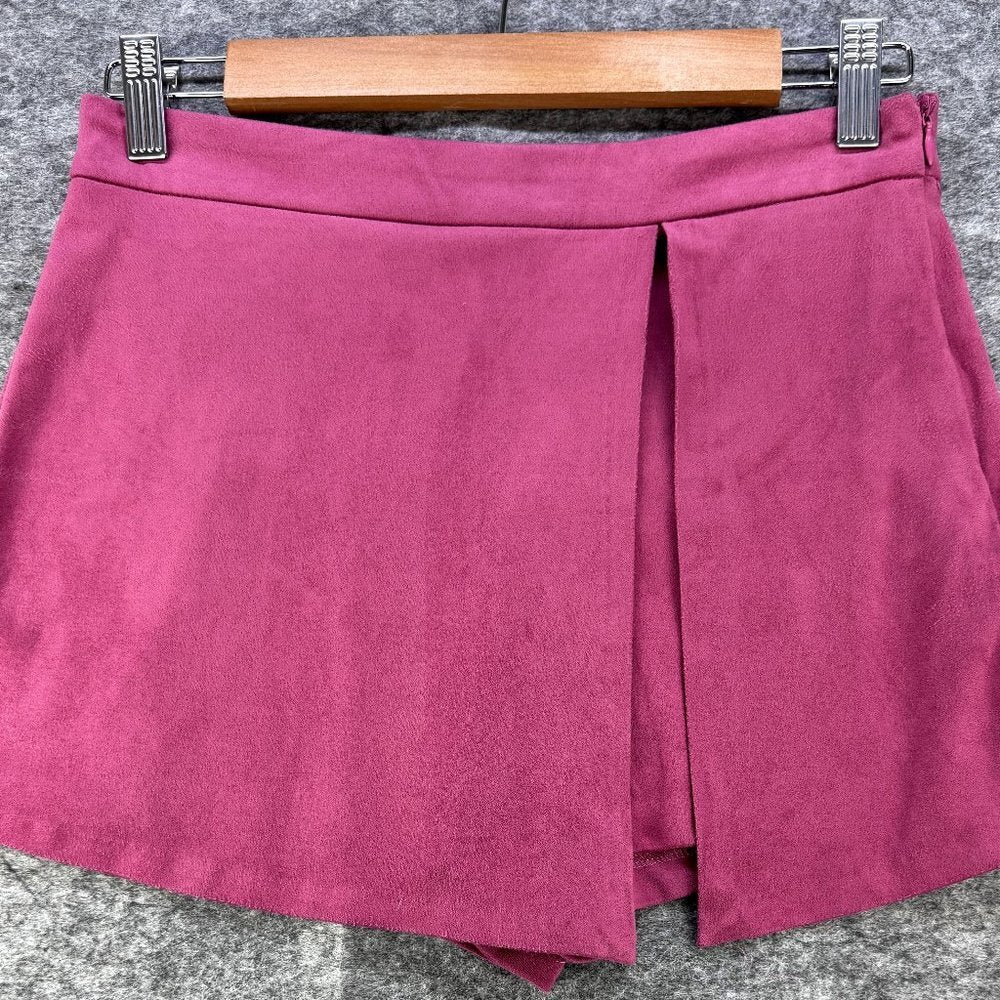 A magenta pink faux suede wrap mini skirt with a front slit, hanging from a wooden hanger with metal clips against a gray textured background.