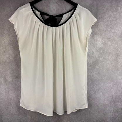 An off-white or cream-colored sleeveless top with a black trim around the scoop neckline, hanging on a black hanger against a textured gray background.