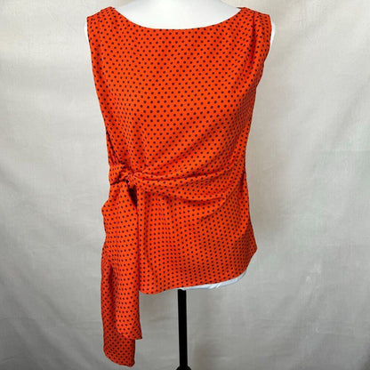 An orange sleeveless top with a black polka dot pattern, styled on a white mannequin torso with a side tie detail, set against a light gray fabric backdrop.