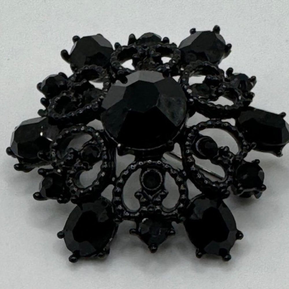 A black ornate brooch featuring a central faceted black gemstone surrounded by smaller black teardrop and oval stones, with intricate filigree detailing, set against a plain light background.