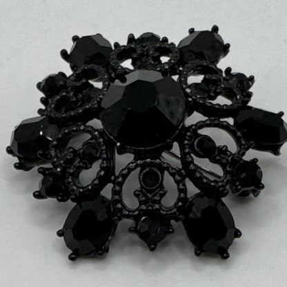 A black ornate brooch featuring a central faceted black gemstone surrounded by smaller black teardrop and oval stones, with intricate filigree detailing, set against a plain light background.