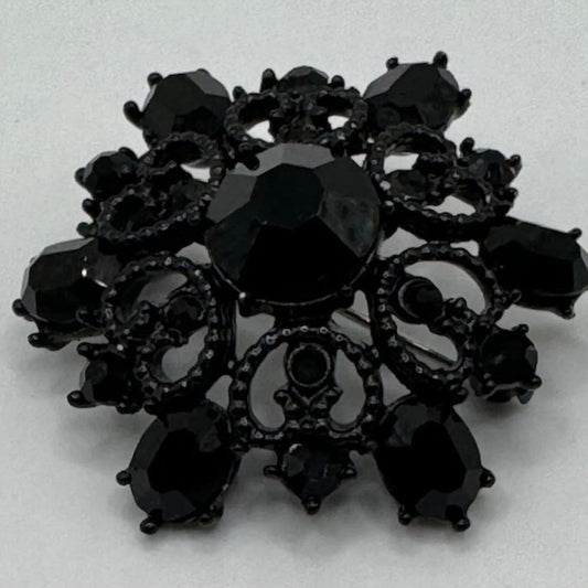 A black ornate brooch featuring a central faceted black gemstone surrounded by smaller black teardrop and oval stones, with intricate filigree detailing, set against a plain light background.