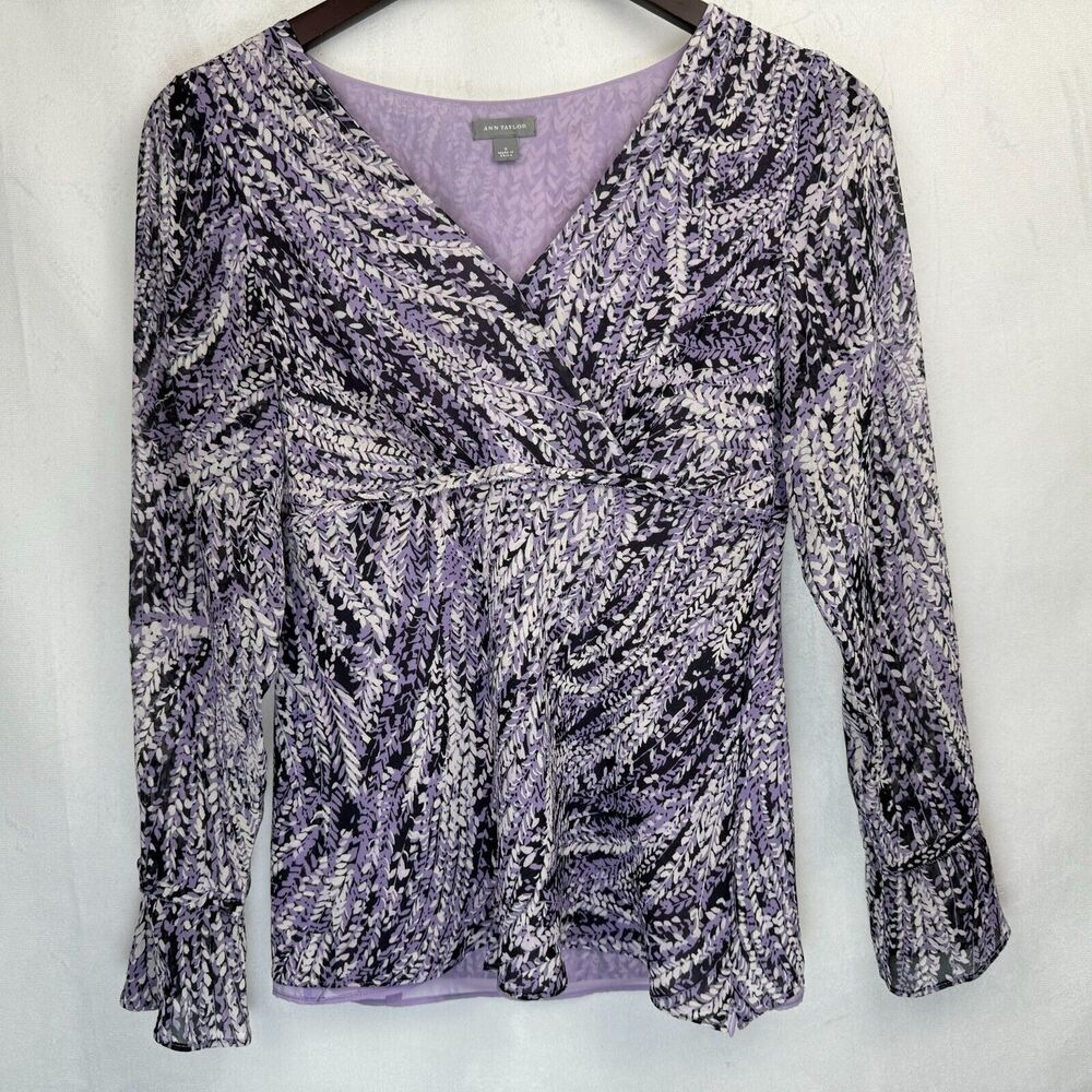 A women's long-sleeve wrap-style blouse with a textured, marled pattern in lavender, black, and white, hanging on a dark wooden hanger against a light gray fabric backdrop.