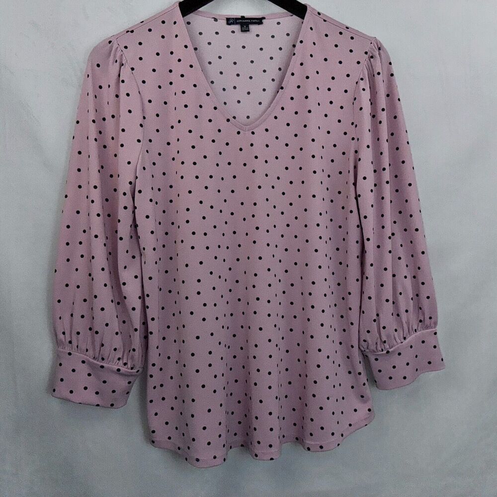 A women's long-sleeve blouse in dusty pink with a black polka dot pattern, featuring a V-neckline and gathered elbow-length sleeves, shown hanging on a black hanger against a light gray fabric backdrop.