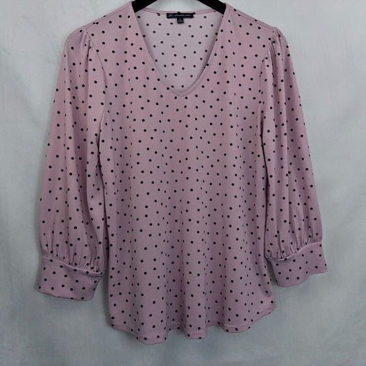 A women's long-sleeve blouse in dusty pink with a black polka dot pattern, featuring a V-neckline and gathered elbow-length sleeves, shown hanging on a black hanger against a light gray fabric backdrop.