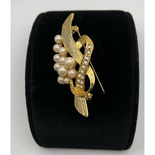 A gold-tone abstract pin featuring curved leaf-like shapes, a cluster of faux pearls, and a vertical section set with small clear rhinestones, displayed on a black velvet ring stand.