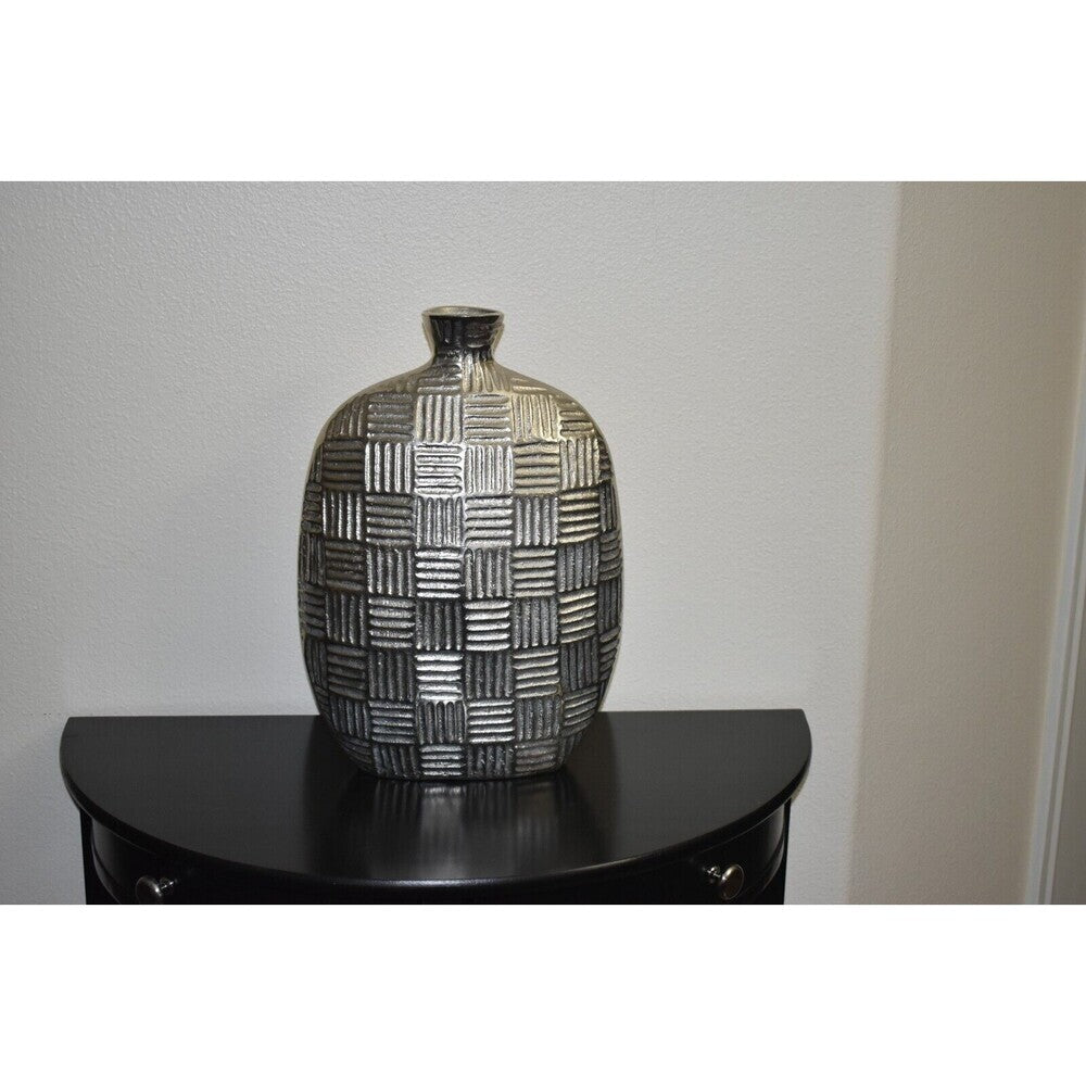 A textured ceramic vase with a metallic silver and black basketweave pattern, displayed on a black curved side table against a light gray wall.