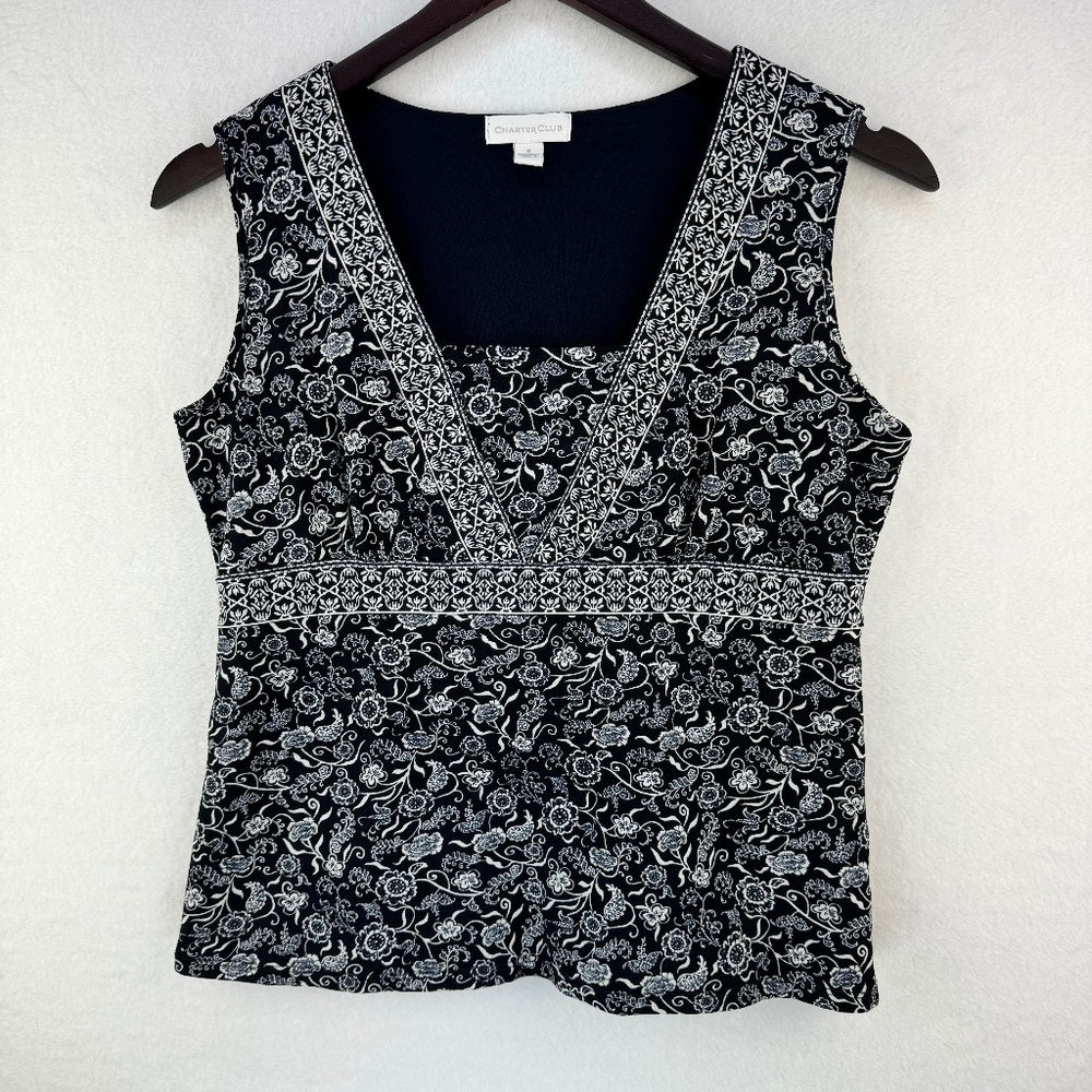A sleeveless black and white floral print top hanging on a dark wooden hanger against a textured white wall. The top features a V-neckline with a solid black inner lining and a decorative horizontal band separating the upper and lower sections.