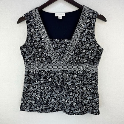 A sleeveless black and white floral print top hanging on a dark wooden hanger against a textured white wall. The top features a V-neckline with a solid black inner lining and a decorative horizontal band separating the upper and lower sections.
