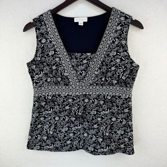 A sleeveless black and white floral print top hanging on a dark wooden hanger against a textured white wall. The top features a V-neckline with a solid black inner lining and a decorative horizontal band separating the upper and lower sections.