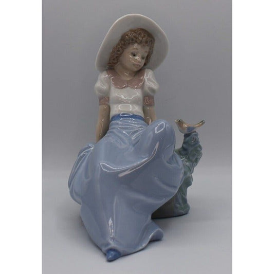 A vintage Lado figurine of a young girl in a white dress and wide-brimmed hat, sitting on a blue-green tree stump with a small bird perched on it, set against a neutral gray background.