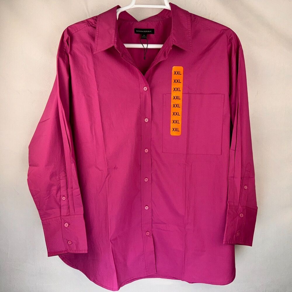 A magenta long-sleeve button-up shirt hanging on a white hanger against a light beige background. The shirt features a vertical orange size tag on the chest pocket listing 'XXXL'.