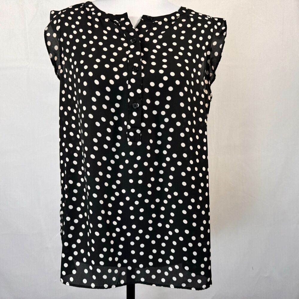 A sleeveless black blouse with an all-over white polka dot pattern, featuring a button-down front and ruffled shoulder details, displayed on a mannequin torso against a white fabric backdrop.