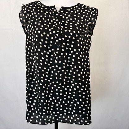 A sleeveless black blouse with an all-over white polka dot pattern, featuring a button-down front and ruffled shoulder details, displayed on a mannequin torso against a white fabric backdrop.