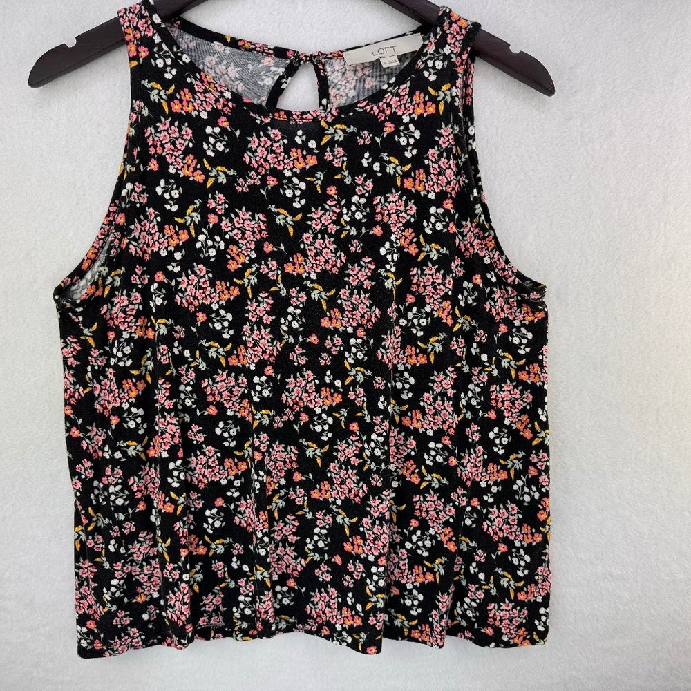 A sleeveless black tank top with a dense pink, orange, and white floral print, hanging on a dark wooden hanger against a textured white wall.