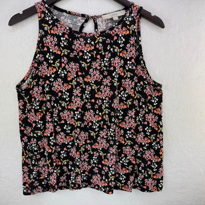 A sleeveless black tank top with a dense pink, orange, and white floral print, hanging on a dark wooden hanger against a textured white wall.