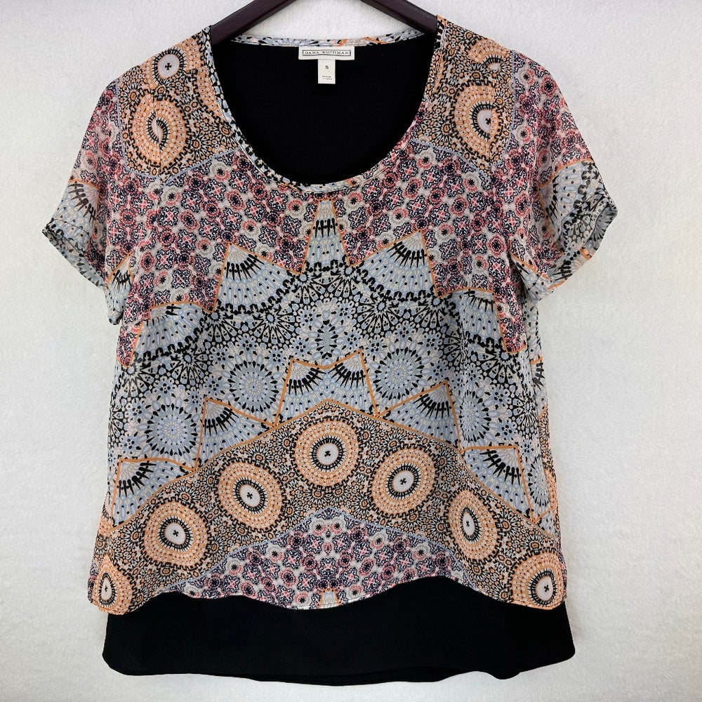 A short-sleeve blouse with a complex geometric and floral mandala pattern in black, white, orange, and purple, hanging on a black hanger against a light textured wall.
