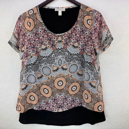 A short-sleeve blouse with a complex geometric and floral mandala pattern in black, white, orange, and purple, hanging on a black hanger against a light textured wall.