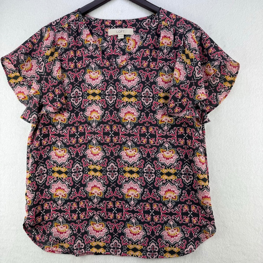 A short-sleeve blouse with a colorful paisley and floral pattern on a black background, hanging on a black hanger against a white textured wall.